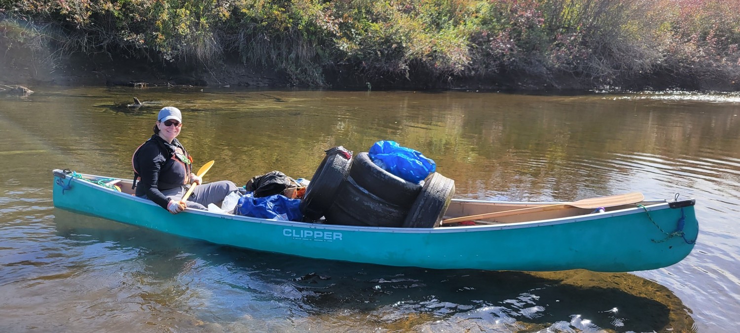 Rebecca Helps (the author) a green canoe filled with collected trash, including tires and bags, as part of a river cleanup effort on a calm and scenic river surrounded by lush foliage.